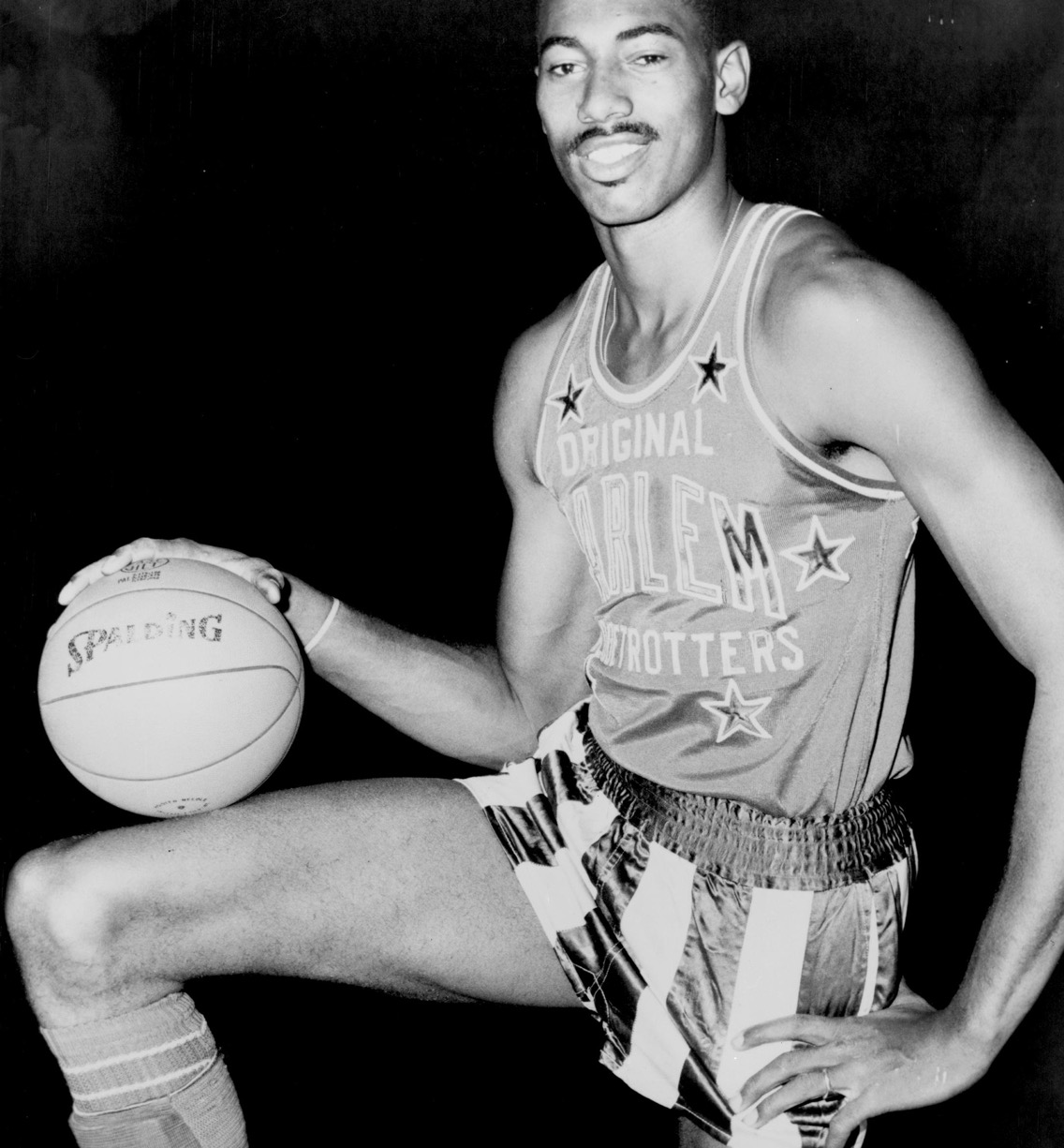 A young Wilt Chamberlain smiling in his Harlem Globetrotters uniform, one hand resting on a basketball balanced on his raised knee