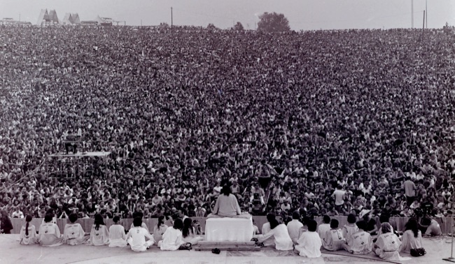 A vast crowd fills a hillside as Swami Satchidananda, seated on a small white-draped stage, leads Woodstock's opening ceremony, 1969