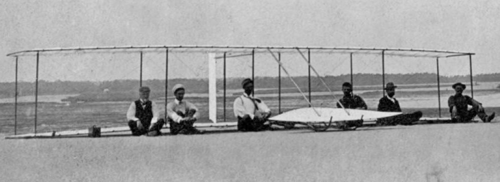 Six men in period clothing seated on the sand in front of an early wood-and-fabric glider at Kitty Hawk in 1902